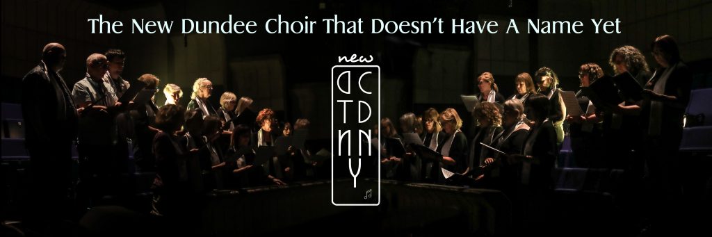 The New Dundee Choir That Doesn't Have A Name Yet singing in Dundee Rep Theatre. The picture is high contrast and dramatic the choir members highlighted against a dark background. 